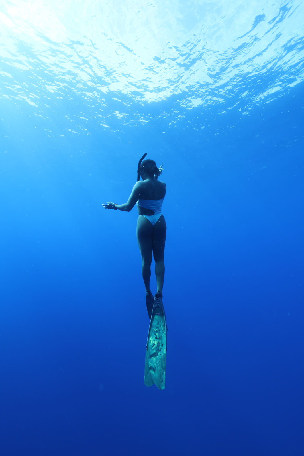 A girl freediving in the pacific ocean in hawaii, wearing long freedving fins and a one piece bathing suit. Items necessary in an adventure travel packing list.