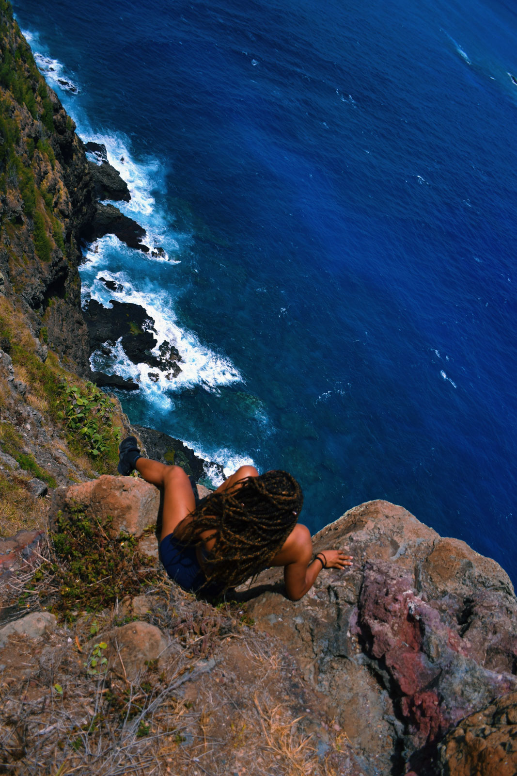 A girl sitting on the edge of a mountain overlooking the ocean in Hawaii. She is wearing sneakers, an item necessary in an adventure travel packing list.