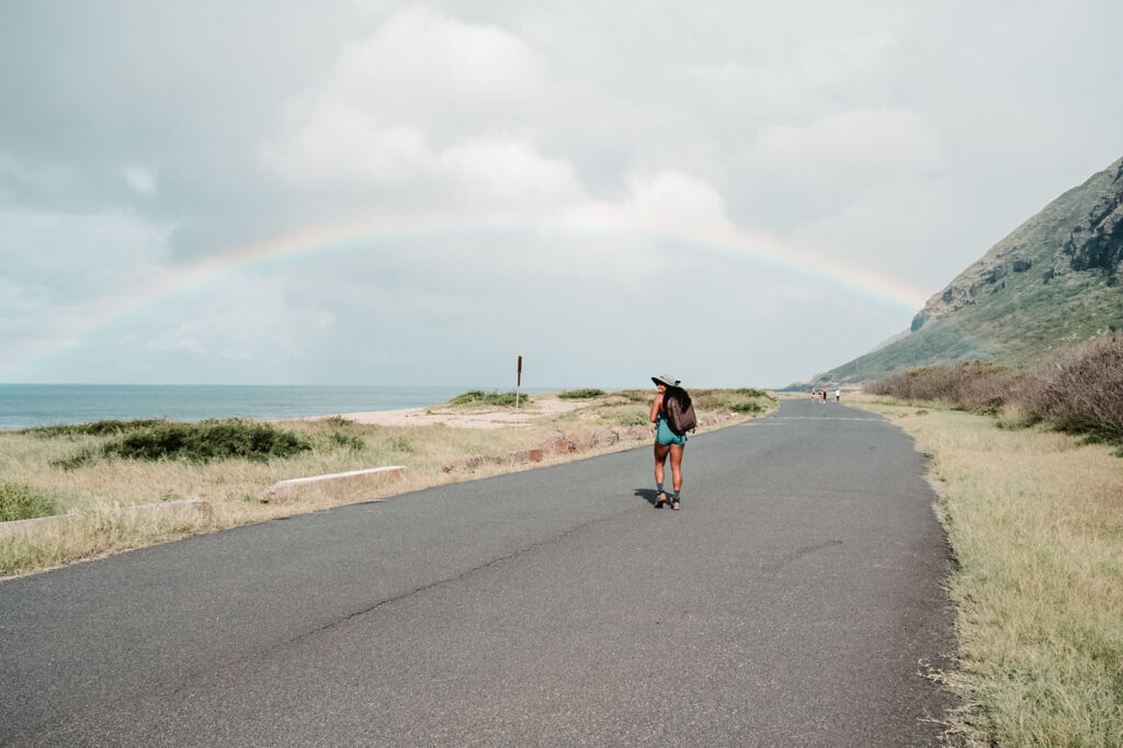 A girl hiking under a rainbow in Hawaii at Kaena point. She's wearing a wide brim hat, shorts and carrying a photography backpack.