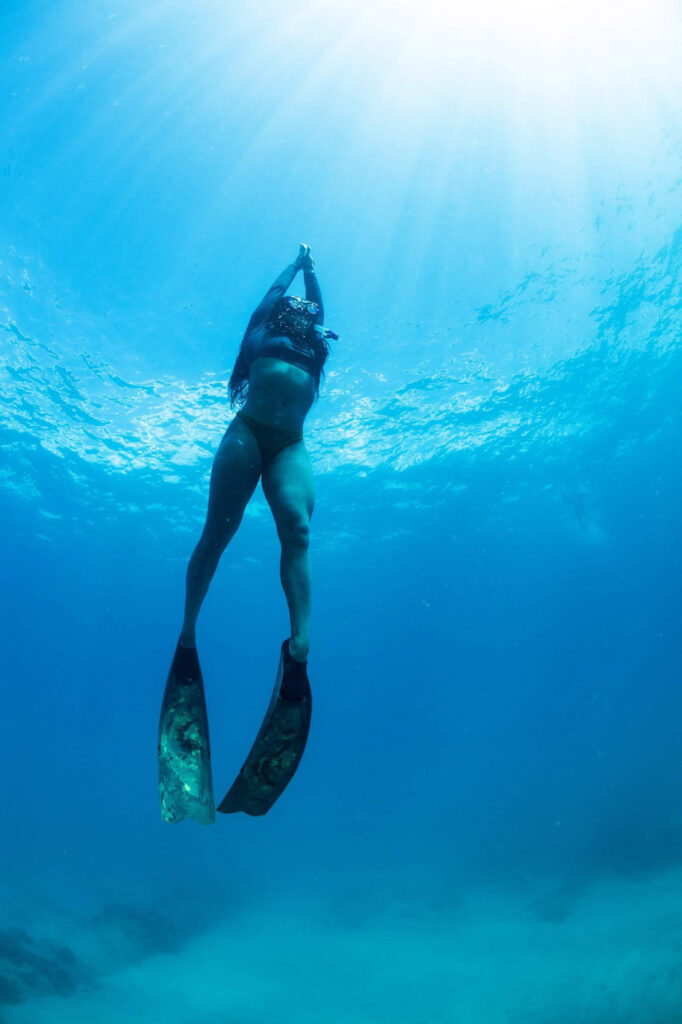 A woman ascending from the bottom of the ocean with her hands pointing towards the surface. She is wearing long fins and a long sleeve rash guard. 