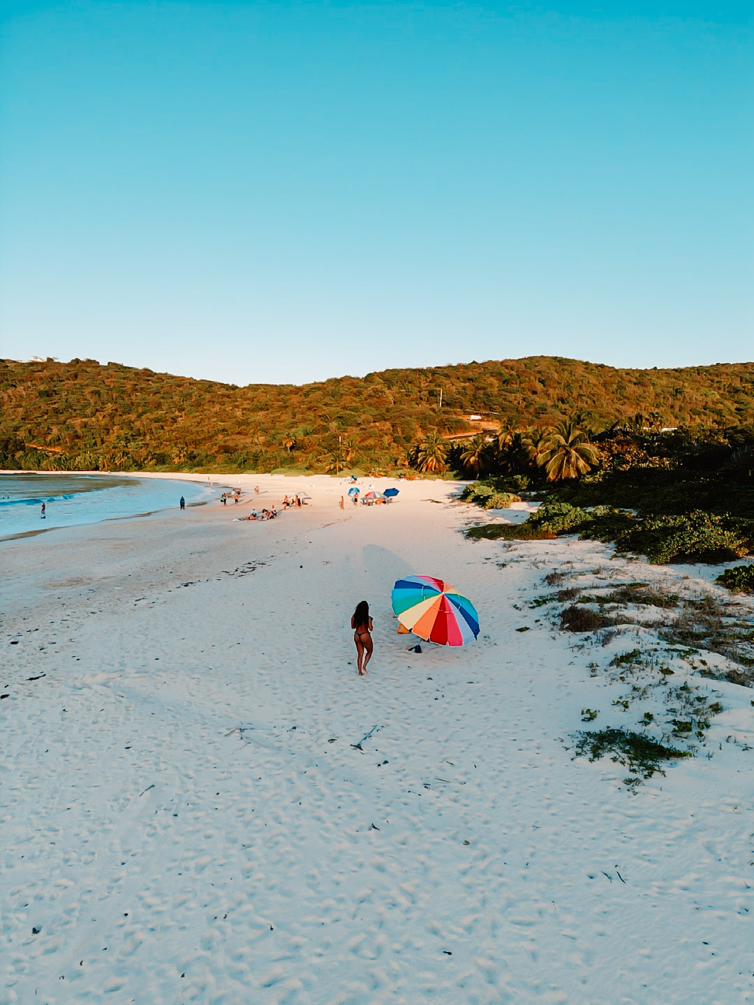 Woman walking on white sand beach in Puerto Rico toward a rainbow beach umbrella at sunset

