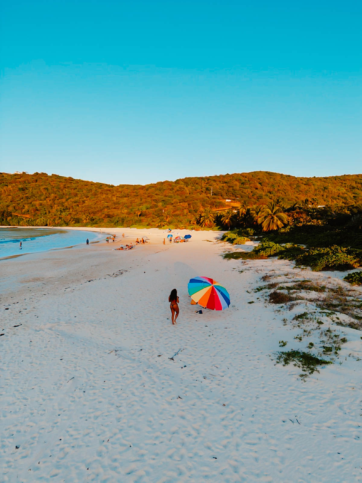 Woman walking on white sand beach in Puerto Rico toward a rainbow beach umbrella at sunset

